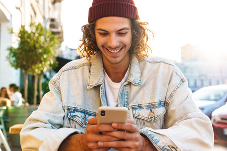 Image of positive young curly man sitting in cafe outdoors and using mobile phone while holding credit cardの写真素材