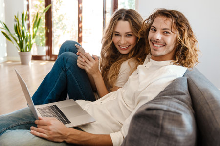 Joyful beautiful couple using cellphone and laptop while resting on couch at homeの写真素材