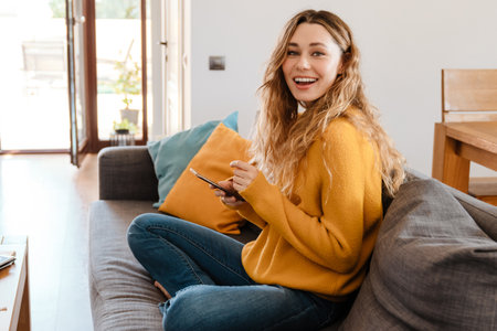 Happy beautiful girl smiling and using cellphone while resting on couch at homeの写真素材