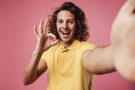Handsome happy young man taking a selfie on a pink background, ok gesture