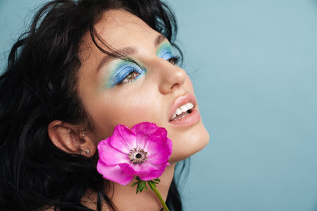 Young beautiful woman wearing bright makeup posing with a flower isolated over blue backgroundの写真素材