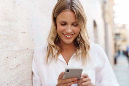 Beautiful happy woman smiling and using cellphone while leaning on wall at city streetの写真素材