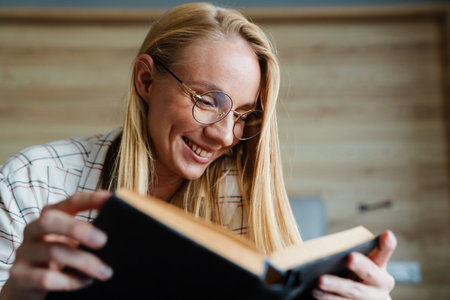 Beautiful blonde pleased woman in eyeglasses reading book while lying in bed at homeの写真素材