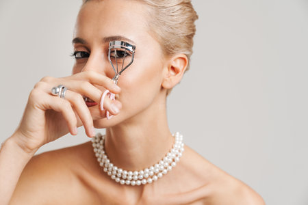 Sensual shirtless girl wearing necklace using eyelash curler isolated over grey backgroundの写真素材
