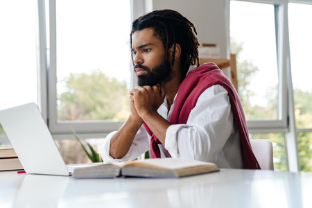 Focused african american guy working with laptop and book while sitting in office