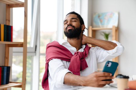 Tired african american guy rubbing his neck and using cellphone while working in officeの写真素材