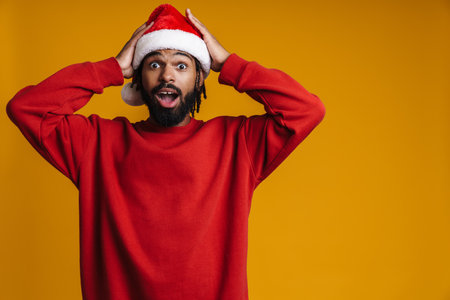 Excited african american guy in Santa Claus hat grabbing his head isolated over yellow backgroundの写真素材