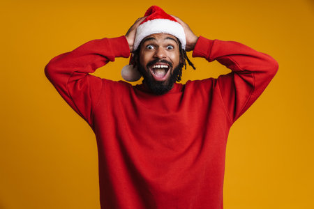 Excited african american guy in Santa Claus hat grabbing his head isolated over yellow backgroundの写真素材