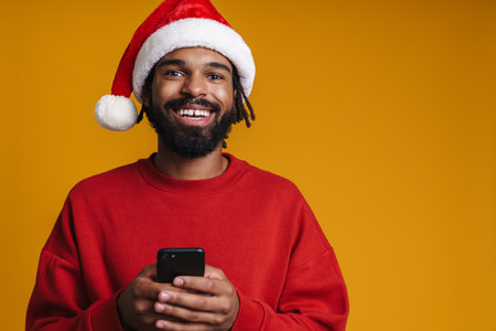 Happy african american guy in Santa Claus hat using cellphone isolated over yellow backgroundの写真素材