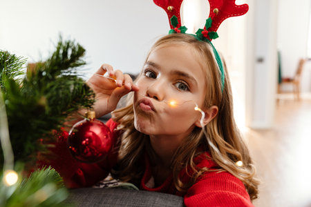 Amusing caucasian girl wearing toy deer horns smiling and holding Christmas lights at homeの写真素材