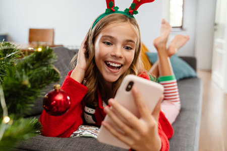 Happy caucasian girl wearing toy deer horns smiling and using cellphone at homeの写真素材