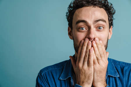 Caucasian excited guy posing and covering his mouth isolated over blue background