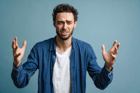 Unhappy handsome guy posing with hands up on camera isolated over blue background