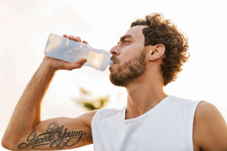 Handsome unshaven guy drinking water while working out on promenade