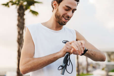 Pleased unshaven guy looking at wristwatch while working out with jump rope on promenade