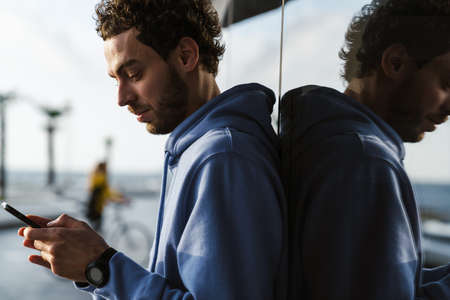 Focused handsome guy using cellphone while leaning on glass wall at city street