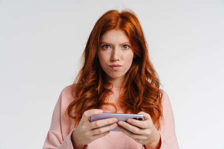 Ginger beautiful displeased girl playing online game on cellphone isolated over white background