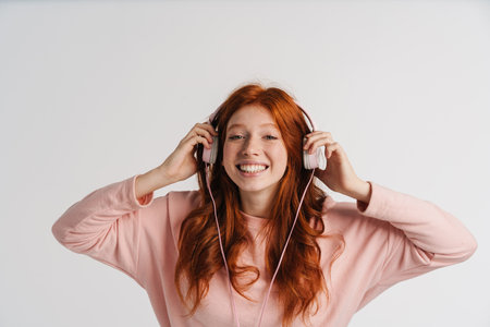 Ginger beautiful happy girl listening music with headphones isolated over white backgroundの写真素材