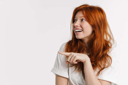 Ginger beautiful excited girl smiling and pointing finger aside isolated over white background