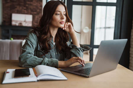 Beautiful focused woman working with laptop while sitting at table indoors