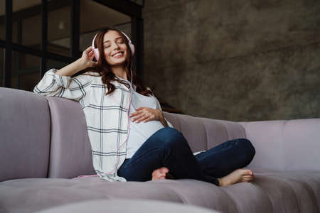 Beautiful happy pregnant woman listening music with headphones while sitting on couch indoors