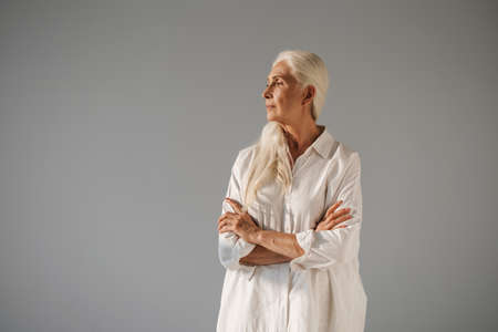 Beautiful elderly white-haired woman posing with arms crossed isolated over grey backgroundの写真素材