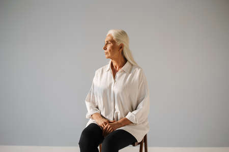 Beautiful elderly white-haired woman looking aside while sitting on chair isolated over grey...