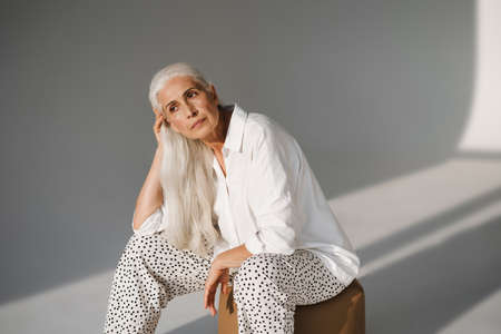 Beautiful elderly white-haired woman looking aside while sitting on chair isolated over grey backgroundの写真素材