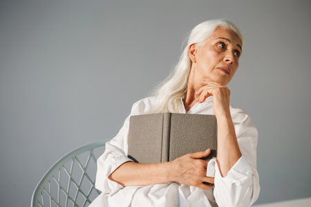 Beautiful elderly white-haired woman sitting with book on chair isolated over grey backgroundの写真素材