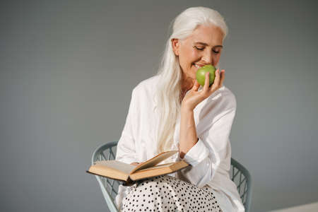 Happy elderly white-haired woman reading book and eating apple isolated over grey backgroundの写真素材