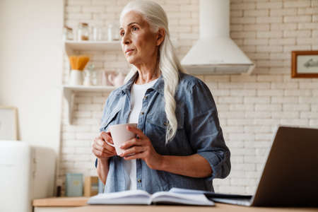 Senior pensive woman working on her laptop in the kitchen, holding cup of teaの写真素材