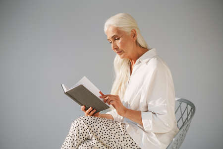 Focused elderly white-haired woman reading book while sitting on chair isolated over grey background