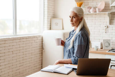 Senior pensive woman working on her laptop in the kitchen, holding cup of tea
