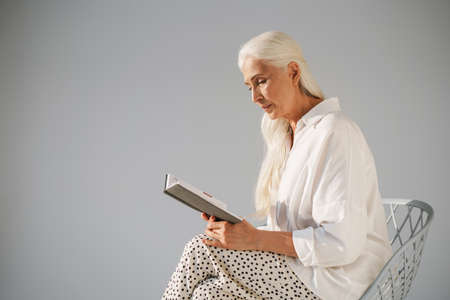 Attractive pensive senior woman reading a book while sitting on a chair isolated over gray backgroundの写真素材