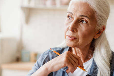 Close up portrait a pensive senior woman thinking indoors, holding pencilの写真素材