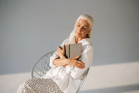 Beautiful elderly white-haired woman sitting with book on chair isolated over grey background