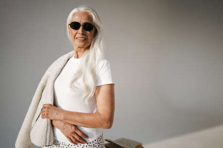 Joyful elderly white-haired woman in sunglasses smiling at camera isolated over grey backgroundの写真素材