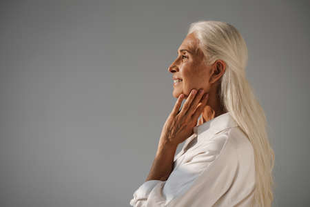 Happy elderly white-haired woman smiling and looking aside isolated over grey background