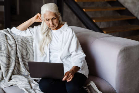 Retired senior woman sitting at home using laptop on a couchの写真素材
