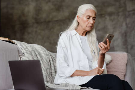 Pensive senior lady sitting on couch at home using cellphone and laptop computer