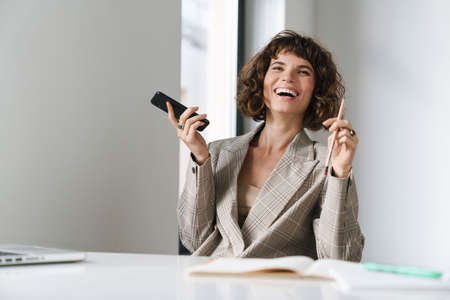 Joyful beautiful girl holding cellphone and making notes while sitting at table in officeの写真素材