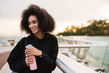 Cheerful athletic sportswoman smiling while standing with water bottle and mat on city bridgeの写真素材