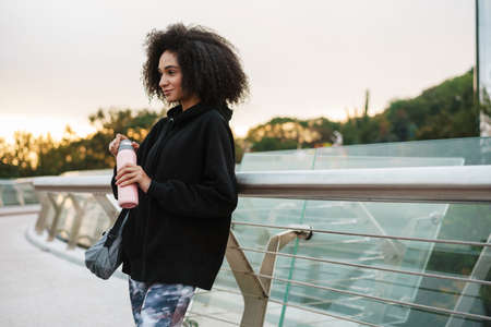 Attractive young african sportswoman carrying fitness mat and holding water bottle walking outdoorsの写真素材