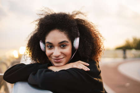 Cheerful nice sportswoman listening music with headphones and smiling on city bridgeの写真素材