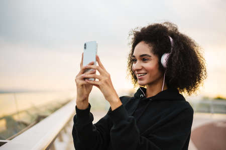 Cheerful athletic sportswoman in headphones taking photo on mobile phone at city bridgeの写真素材