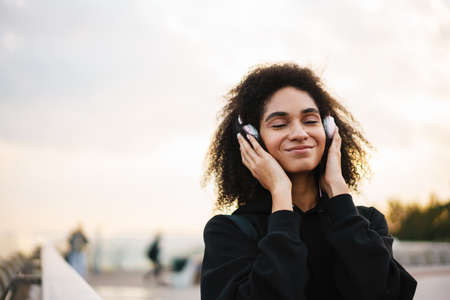 Cheerful nice sportswoman listening music with headphones and smiling on city bridge