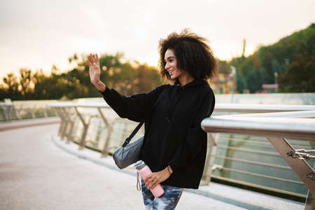 Cheerful sportswoman waving hand while standing with water bottle and mat on city bridgeの写真素材
