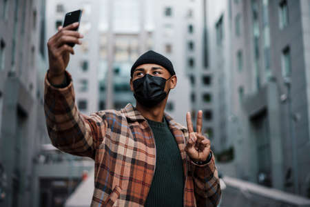 Young afro american man wearing a face mask taking a selfie while walking on a city street