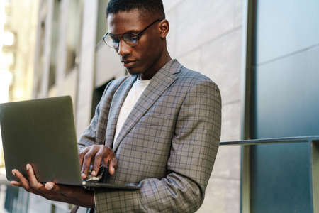Focused african american man smiling and using laptop while standing outdoorsの写真素材