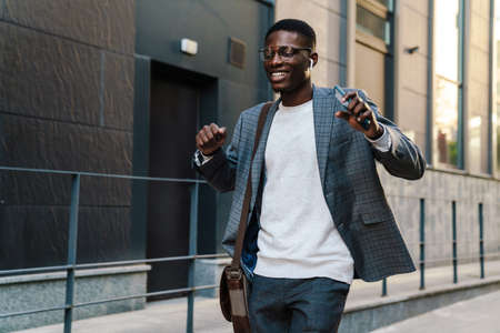 Happy african american man in earphones using mobile phone while dancing on streetの写真素材